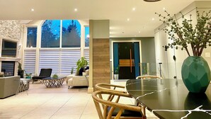 A modern living room with a dining area, large windows, and a view of trees. - Spacious Retreat in the Pines • Walk to Georgian Bay Beaches (Tiny)