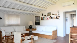 Un salon avec un canapé blanc, deux fauteuils en bois, une petite table avec une lampe, et un mur décoré de tableaux et de livres encadrés.