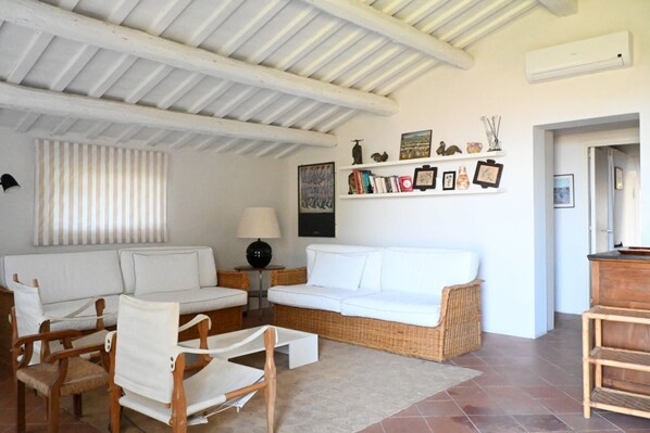 A living room with a white sofa, two wooden chairs, a small table with a lamp, and a wall decorated with framed pictures and books.
