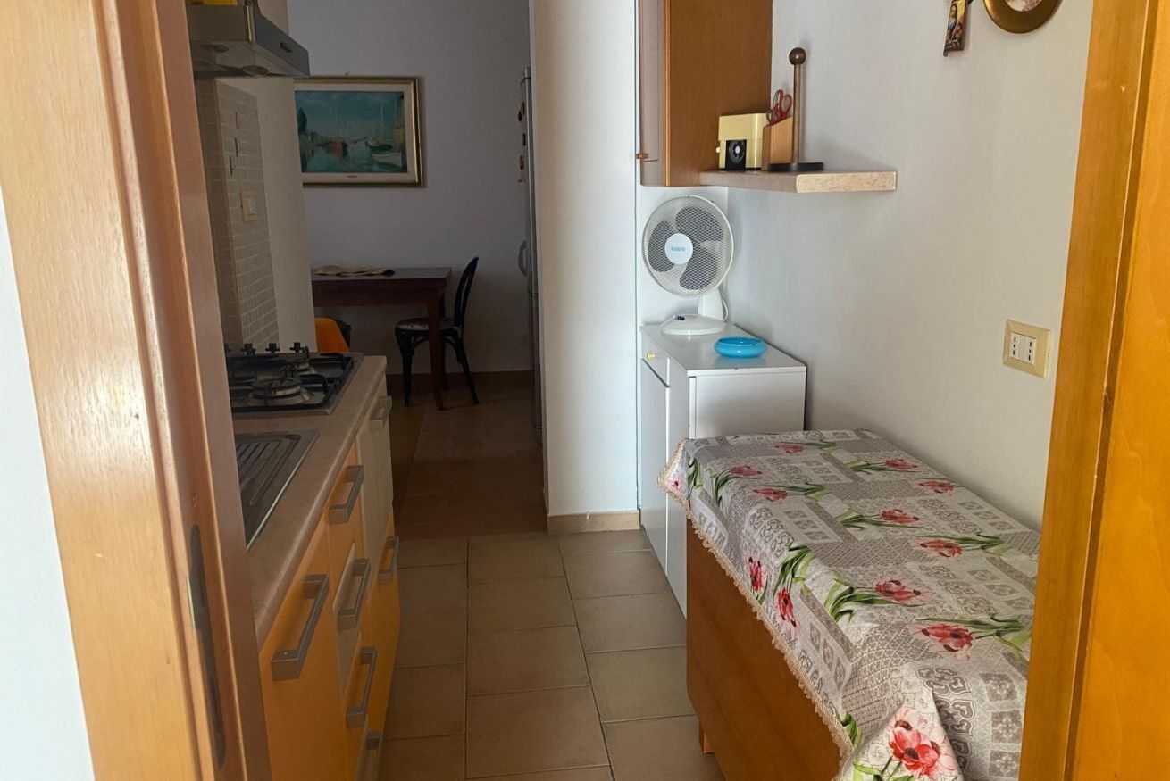 Cocina compacta con estufa a gas, zona de comedor con una silla, una mesita blanca y un ventilador de pared.