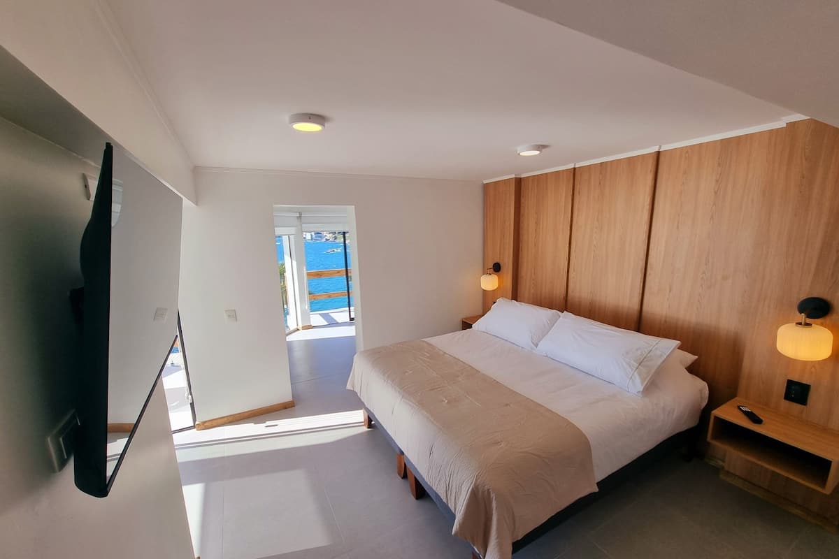 A modern bedroom with a large bed, wooden paneling on the wall, a bedside table with a lamp, and a view of the sea through an open door.