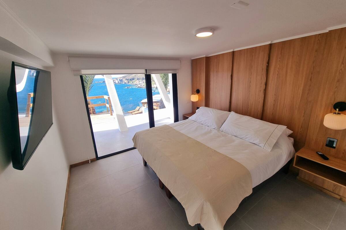 A bedroom with a bed, wooden paneling, a nightstand, a wall-mounted lamp, and a view of the sea through a sliding door.