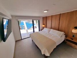 Un dormitorio con cama, paneles de madera, una mesita de noche, una lámpara de pared y vistas al mar a través de una puerta corrediza.