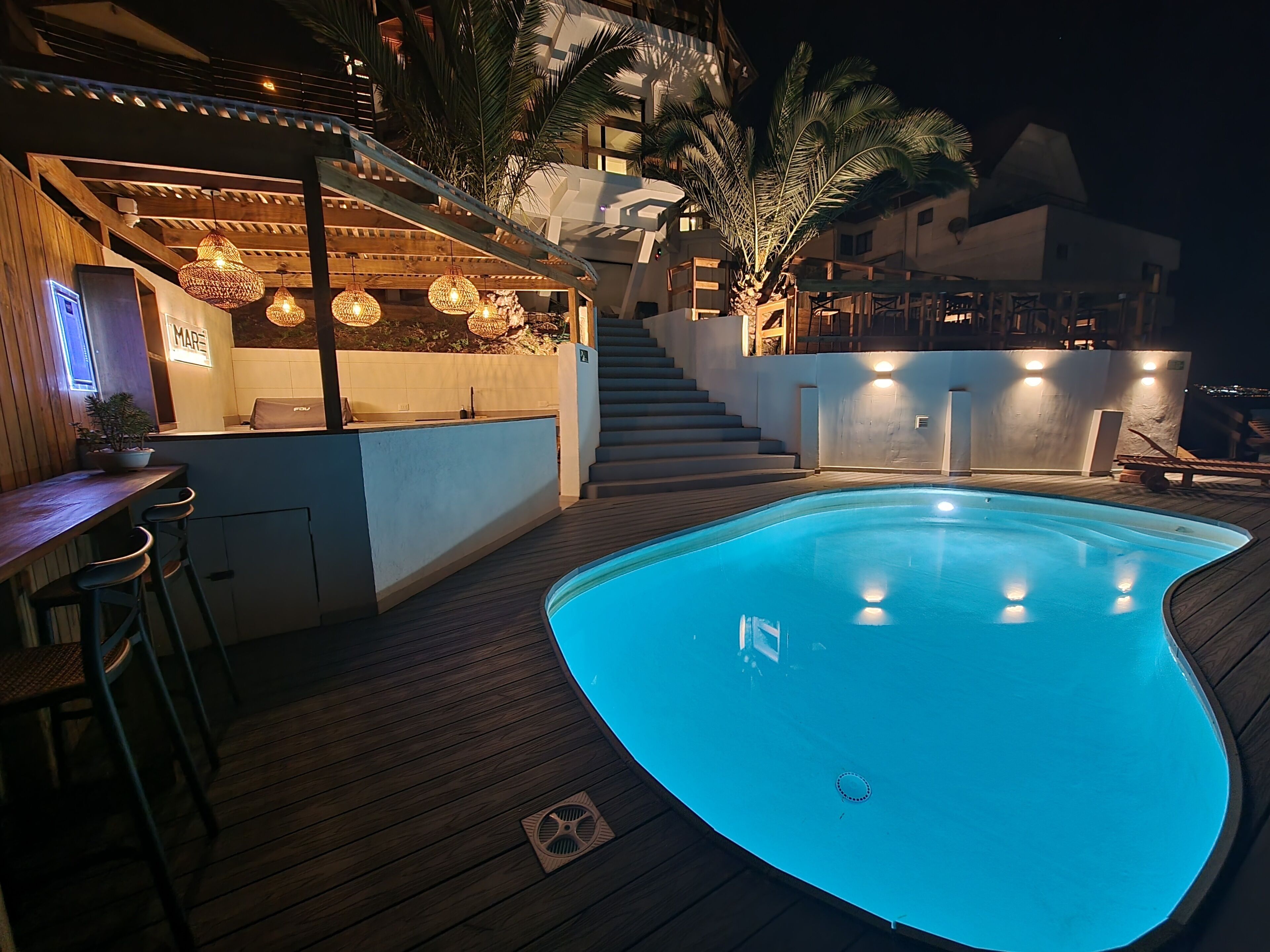 Una piscina con cubierta de madera, una zona de bar con luces colgantes y una escalera que conduce a un nivel superior.