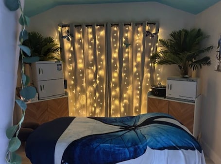 A bedroom with a bed, curtains with string lights, a small cabinet, and a potted plant.