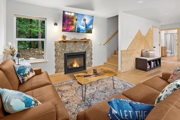 A modern living room with a stone fireplace, a large TV mounted above it, and a cozy seating area with brown sofas and patterned cushions.