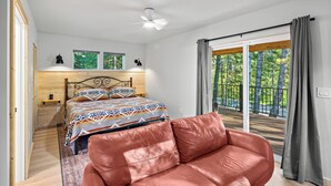 Un dormitorio con cama, un sillón de cuero rojo, cabecera de madera, ventilador de techo y vista a árboles a través de una puerta corrediza de vidrio.