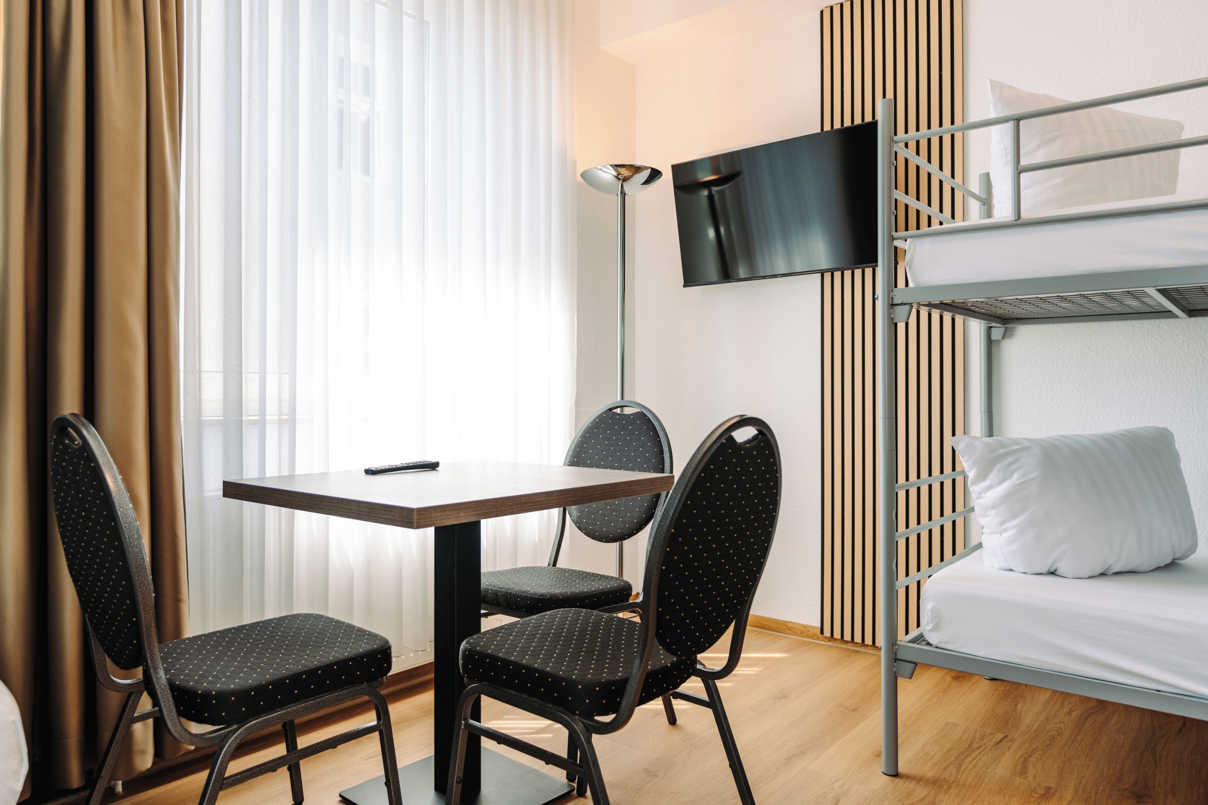 A hotel room with a bunk bed, a desk with two chairs, a television, and a window with curtains.