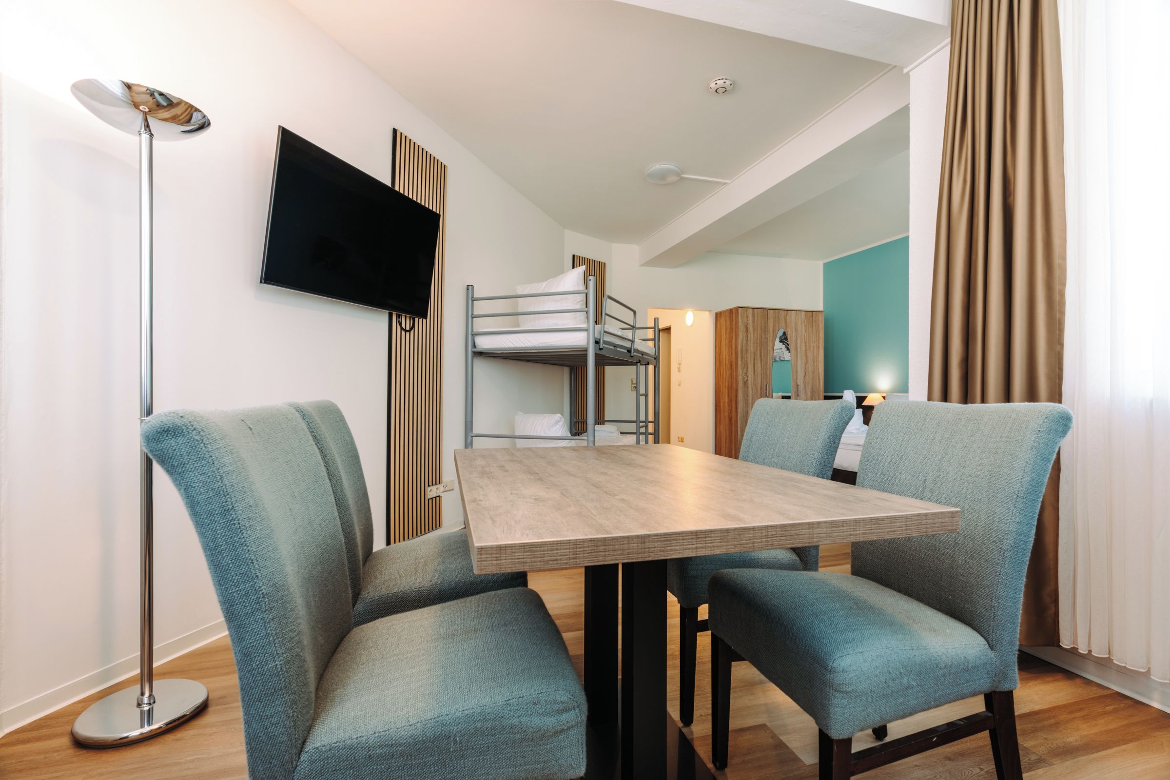 Una habitación de hotel moderna con una cama litera, un televisor de pantalla plana, una mesa de comedor con sillas y una lámpara de pie.