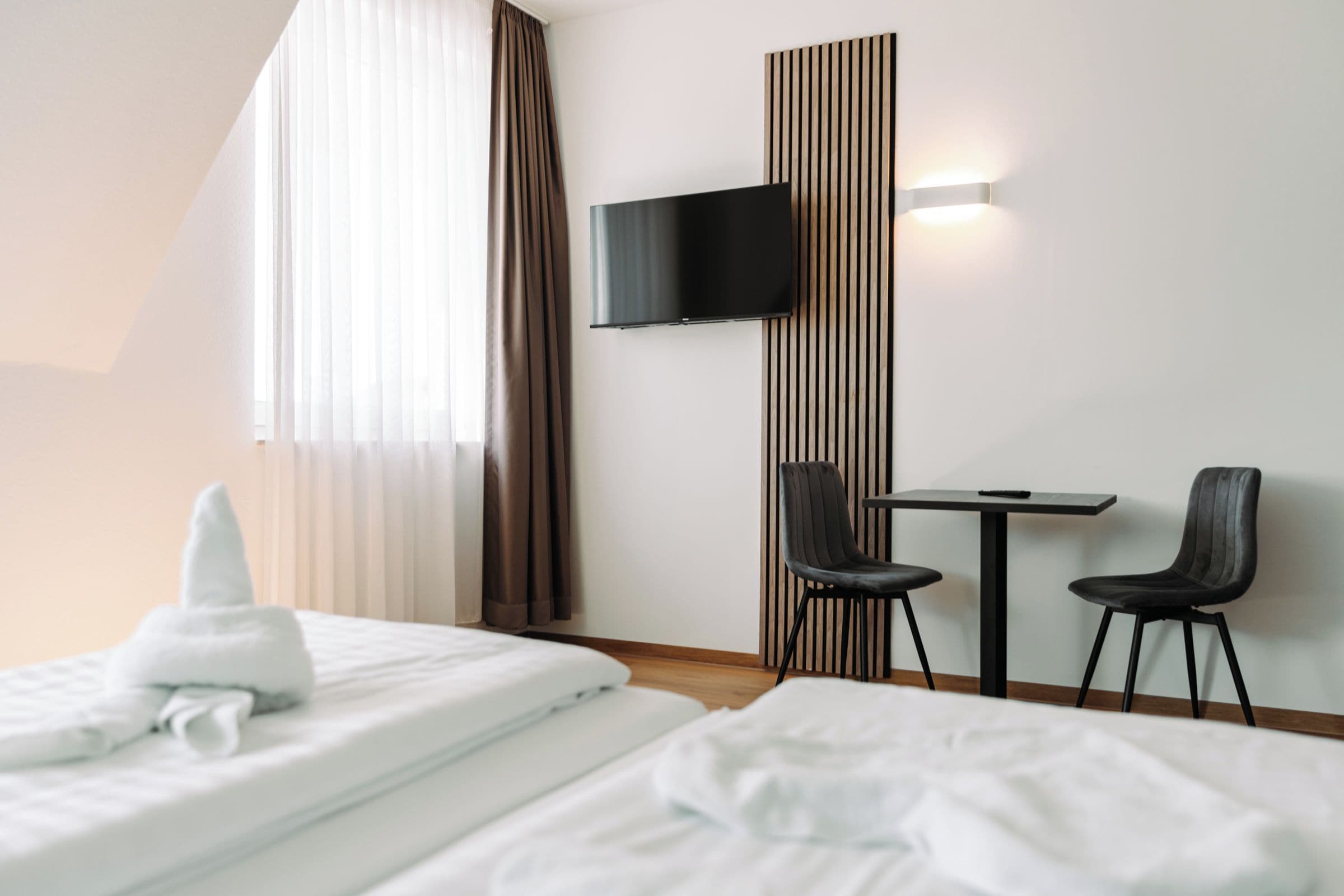 Habitación de hotel con dos camas, un televisor montado en una pared de paneles de madera y una mesita con dos sillas.