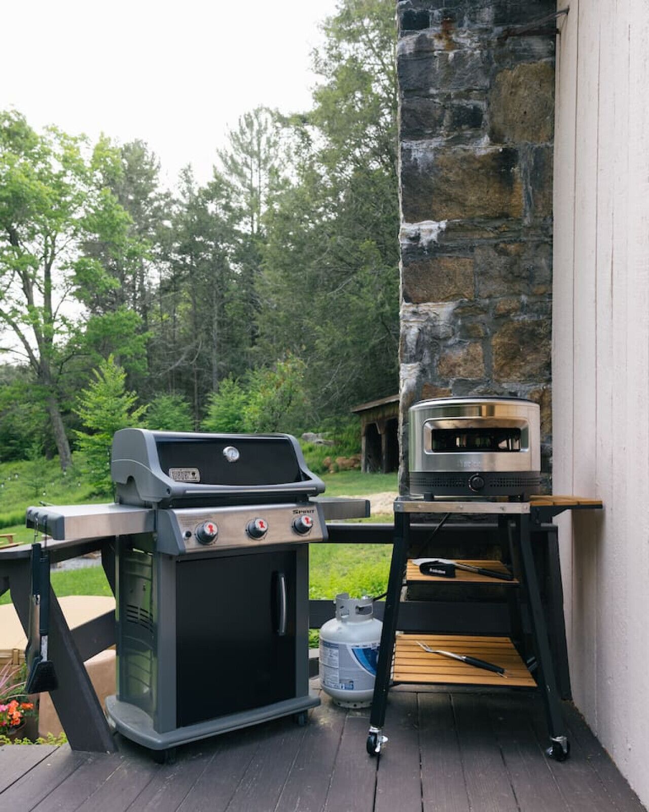Una parrilla exterior y un horno de pizza en una terraza de madera con un muro de piedra de fondo.