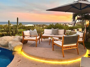 A poolside area with a round table and chairs, a large umbrella, and a view of the ocean and desert landscape.