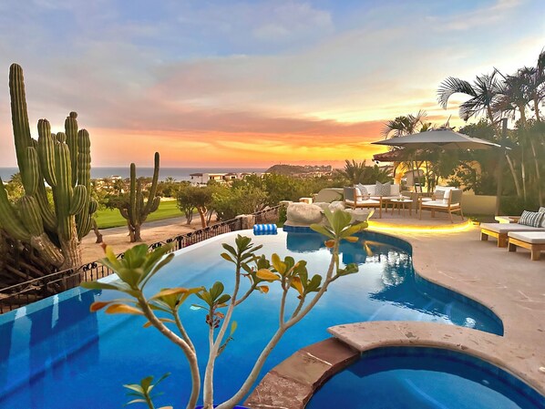 A pool area with a view of the sunset, surrounded by cacti and palm trees.