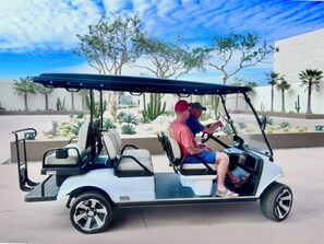 A golf cart with two people, a desert landscape with cacti and palm trees, and a building in the background.