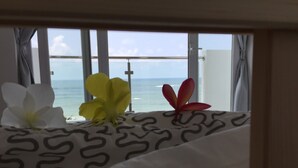A bed with a patterned bedspread and three tropical flowers. A window with curtains reveals a view of the ocean. - Wake up to panoramic ocean views from almost every room. (Dehiwala-Mount Lavinia)