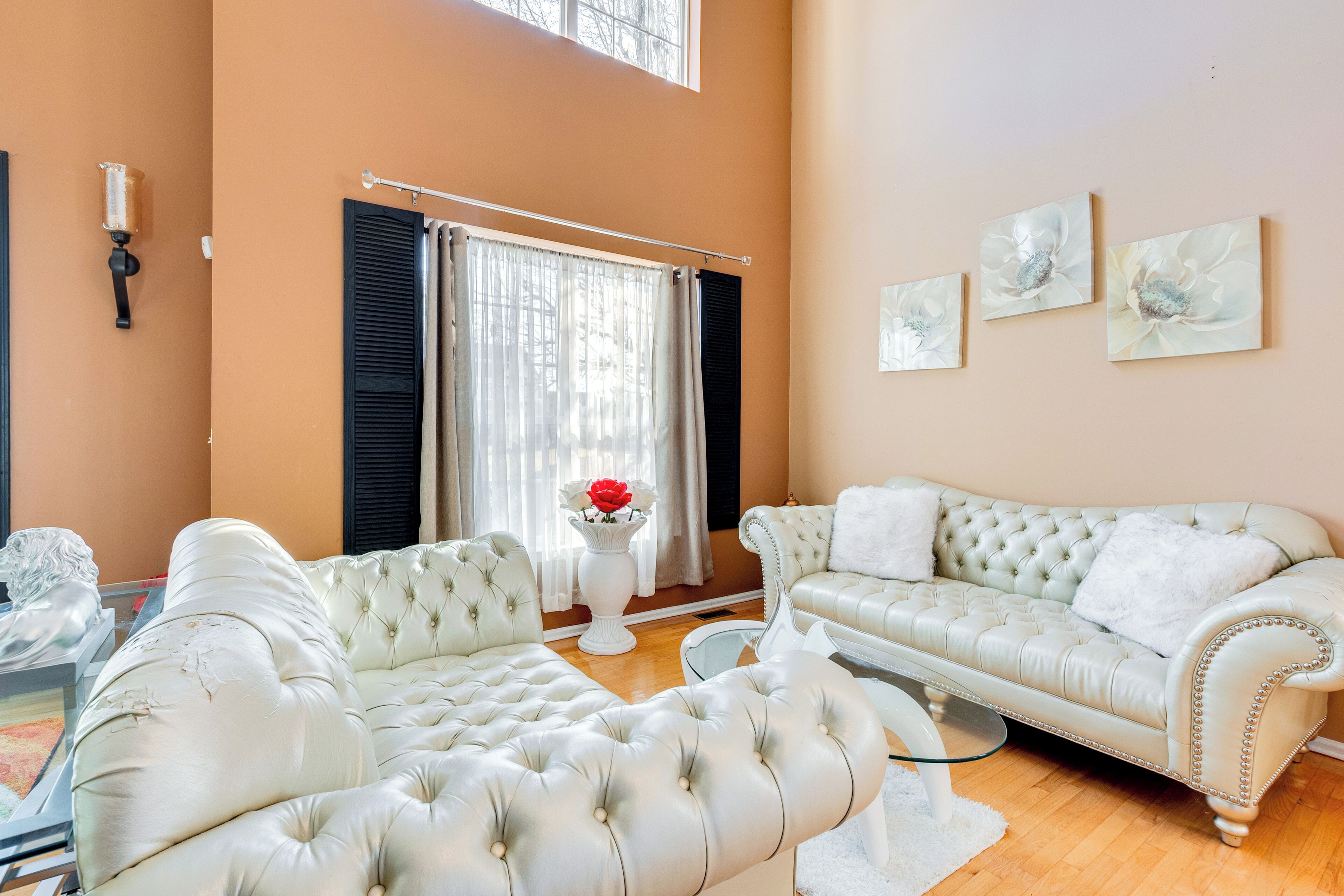 A spacious living room with a large white sofa, a smaller white armchair, and a glass coffee table. There is a window with black shutters and a white vase with red flowers on a pedestal.