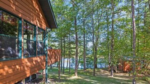 A wooden cabin with large windows, surrounded by a forested area with a lake in the background.