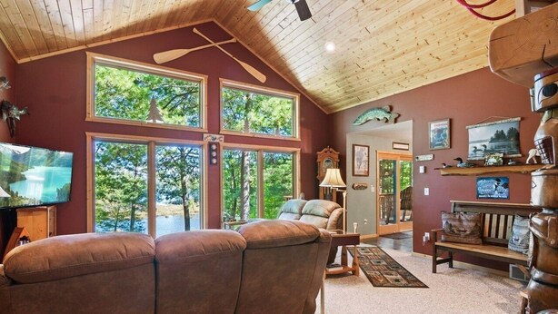 A cozy living room with a large brown sofa, wooden ceiling, and large windows overlooking a natural landscape.