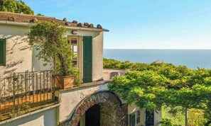 Un edificio costero con balcón, arco de piedra y abundante vegetación.