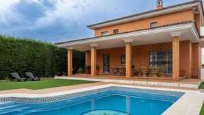 Una piscina con un diseño geométrico, un porche cubierto con mobiliario exterior y una casa de ladrillo con columnas.