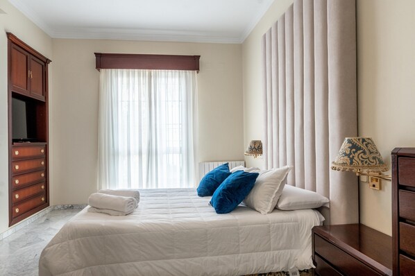 Une chambre d’hôtel avec un lit, une commode, une télévision et une fenêtre avec des rideaux.