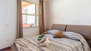 A bedroom with a bed, a window with curtains, a bedside table with a lamp, and a view of the outside.