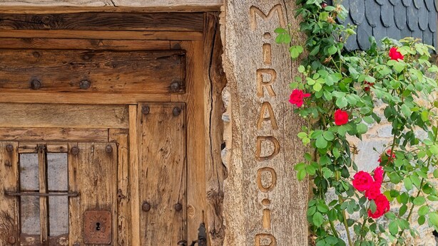 Una puerta de madera rústica con un pestillo de metal y un ventanal pequeño, un muro de piedra con el nombre 'MIRADOR' grabado y rosales rojos creciendo en la pared.