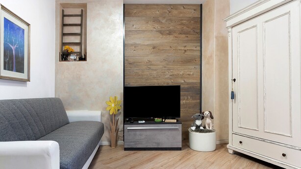 Un salón con un sofá gris, un televisor de pantalla plana sobre un mueble de madera, un armario blanco y un estante con objetos decorativos.