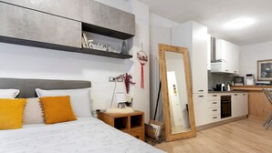 Un dormitorio moderno con cama, mesita de noche y un espejo grande. Junto a él, una cocina con armarios blancos y encimera de madera.