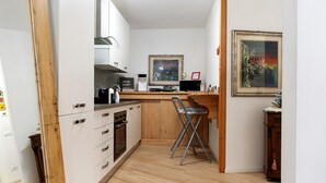 Cocina compacta con armarios blancos, encimera de madera, una silla plegable y un cuadro en la pared.