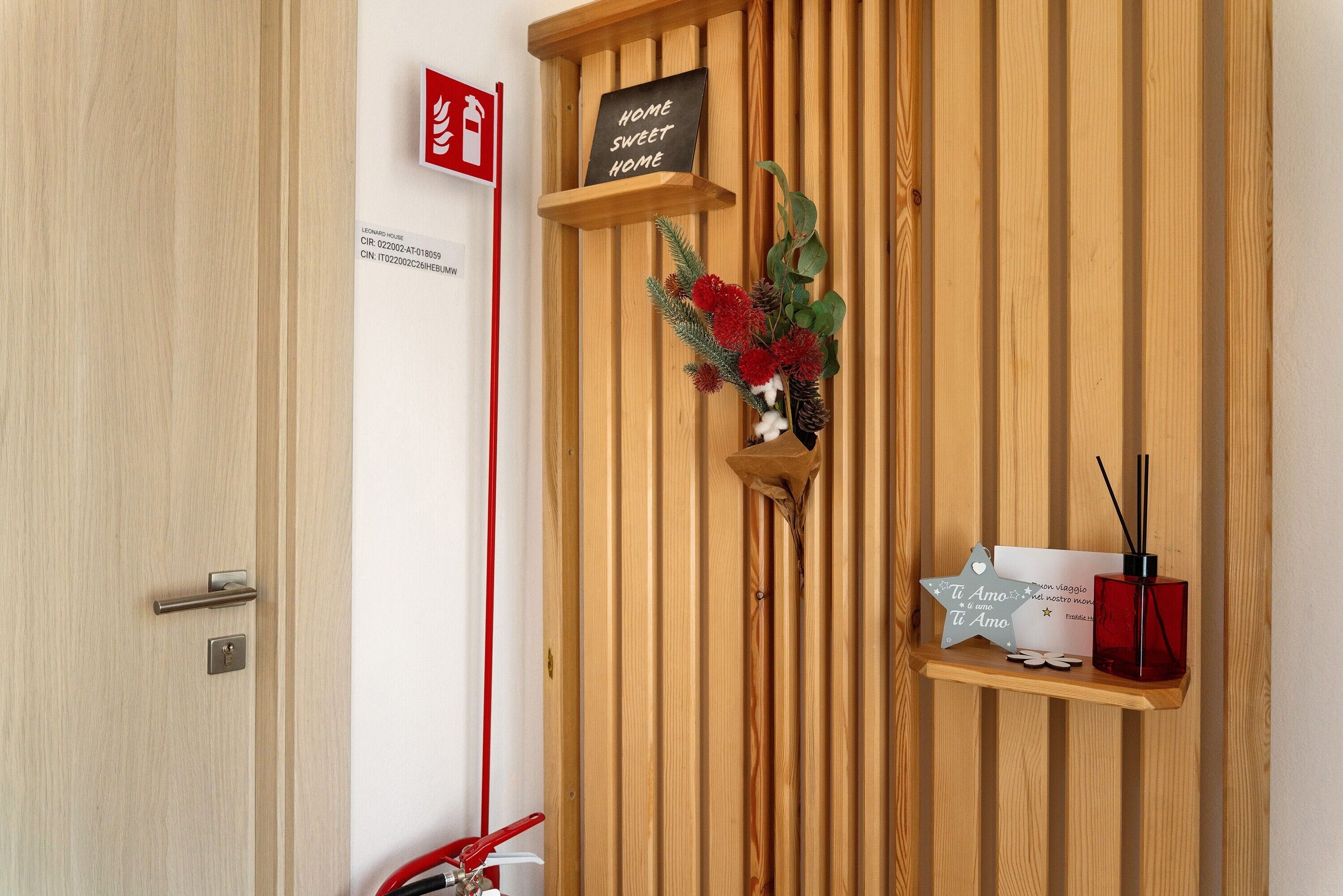 Een houten wand met een bord waarop staat 'HOME SWEET HOME', een klein plankje met een decoratief item en een rood voorwerp op de vloer.