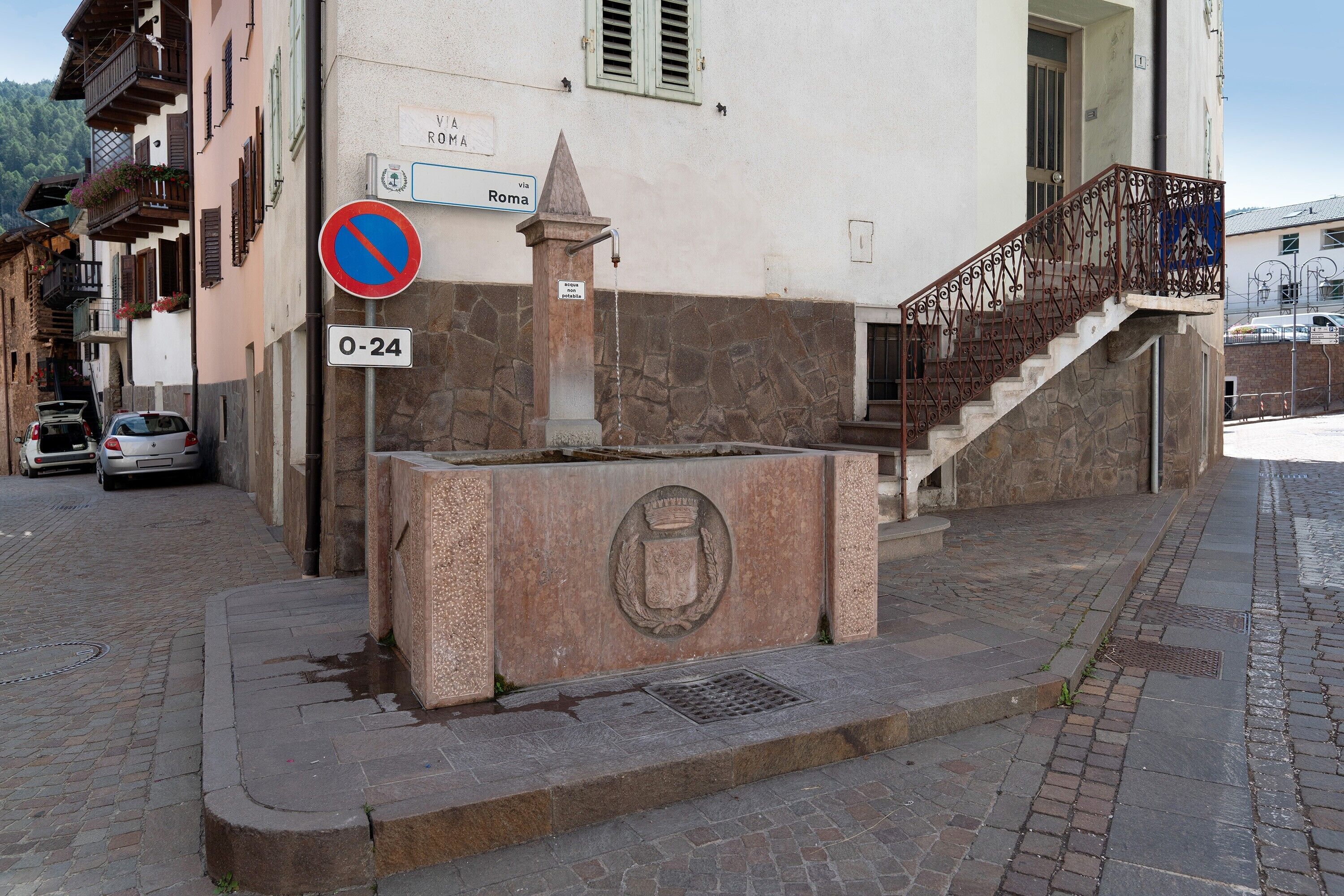 Une rue avec une fontaine en pierre, un panneau indiquant qu’il est interdit de se garer, et un escalier menant à un immeuble.