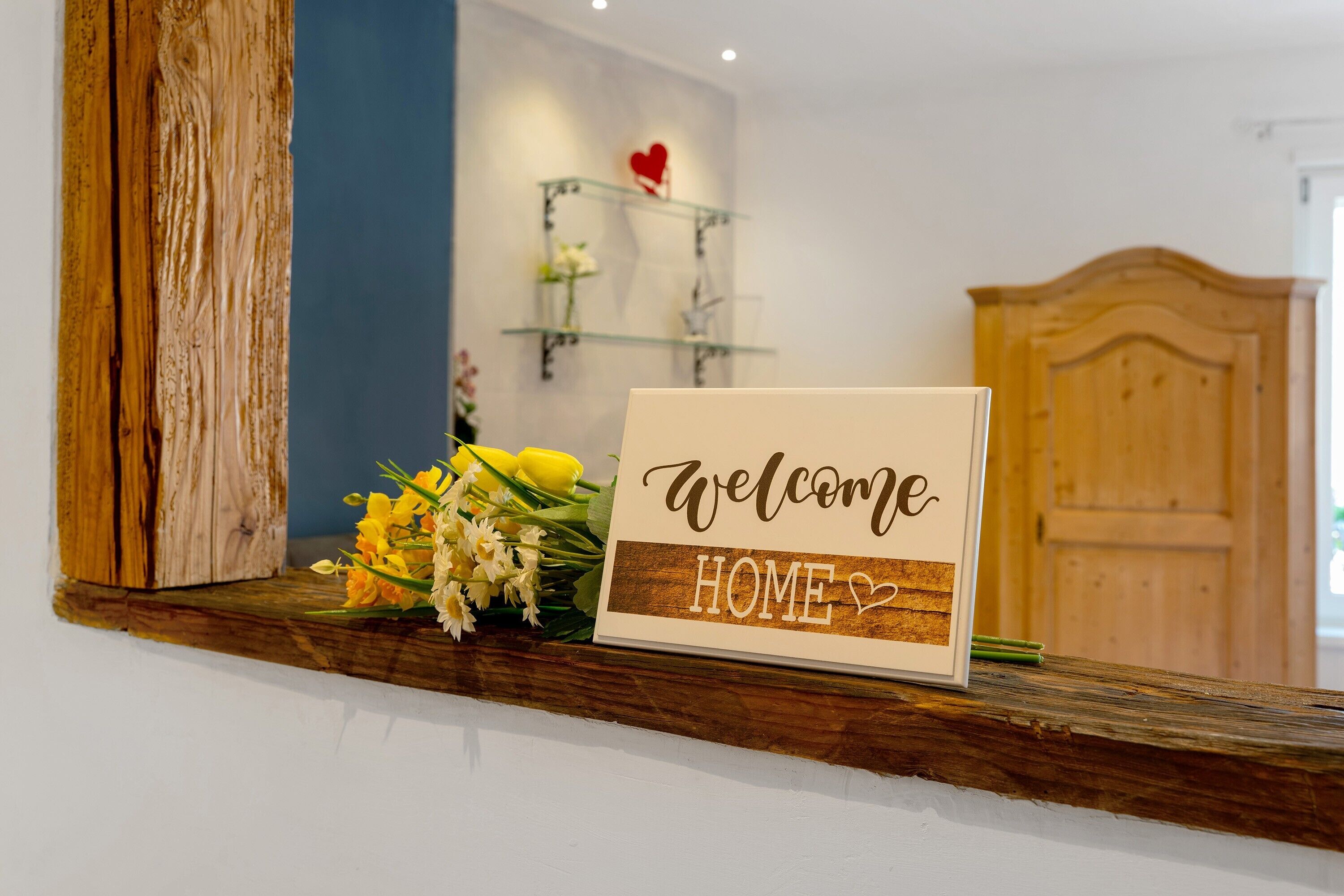 Ein Willkommen-Schild mit der Aufschrift 'WELCOME HOME' auf einem Holzregal, ein Blumenstrauß und im Hintergrund eine Holztür.