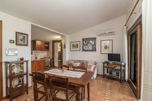 A dining area with a wooden table and chairs, a kitchen in the background, and artwork on the walls.