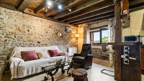 Una habitación rústica con pared de piedra, vigas de madera, un sofá, una silla, una mesa de centro de vidrio, un taburete de madera y una estantería para vinos.