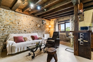 Une pièce rustique avec un mur en pierre, des poutres apparentes, un canapé, un fauteuil, une table basse en verre, un tabouret en bois et un meuble à vin.