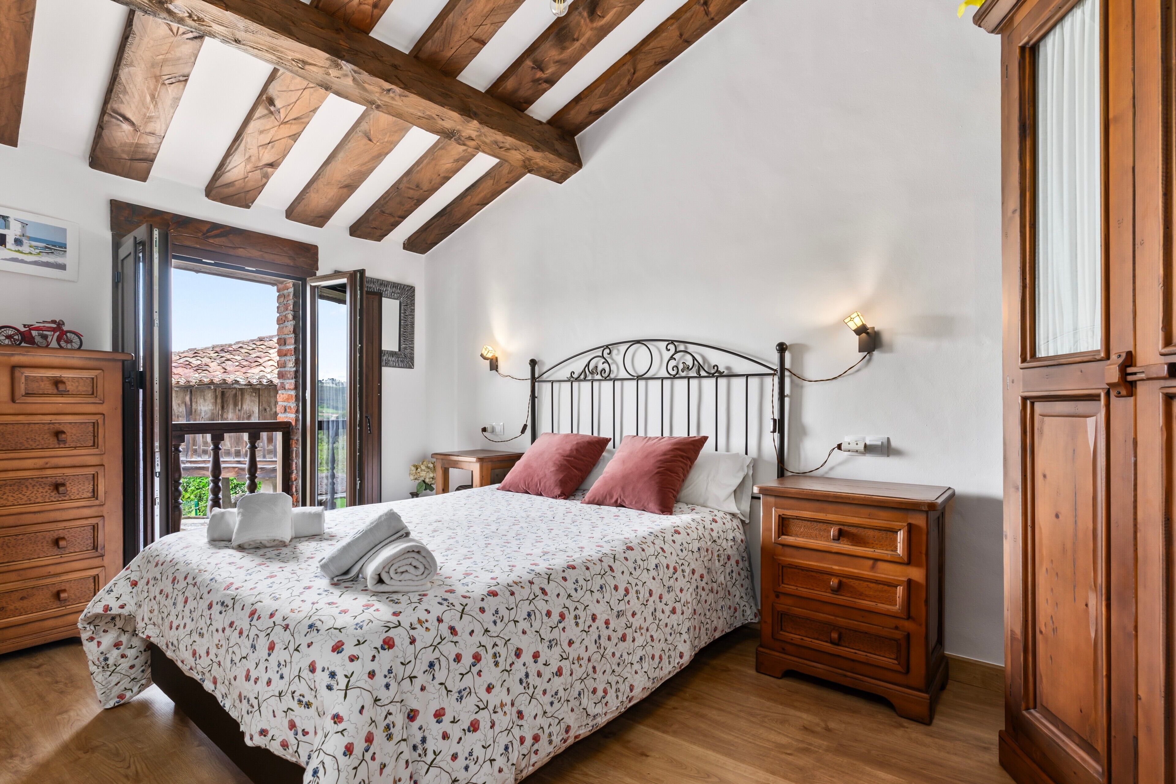 A bedroom with a wooden ceiling, a bed with a floral bedspread, a nightstand, a dresser, and a window with a view of buildings.