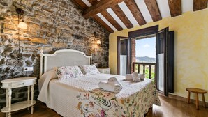 Un dormitorio con pared de piedra, vigas de madera, cama con ropa de cama floral, una mesita de noche y vistas a través de una ventana.