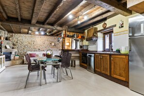 A rustic kitchen with stone walls, wooden beams, and a dining area with a glass table and wicker chairs.
