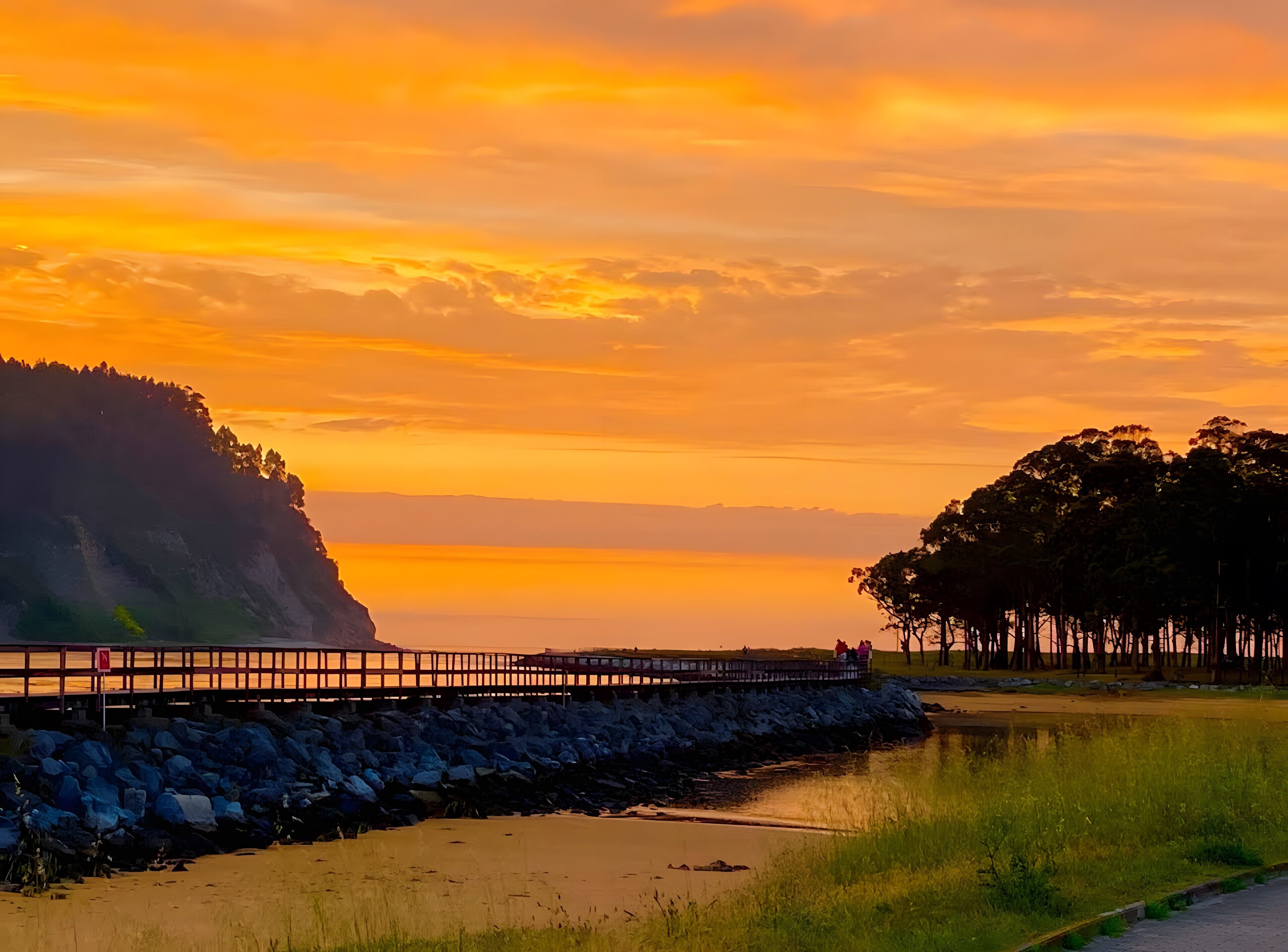 Ein Küsten-Sonnenuntergang mit einem hölzernen Steg, der sich in ruhiges Wasser erstreckt, umgeben von felsigem Gelände und dichtem Baumbestand.