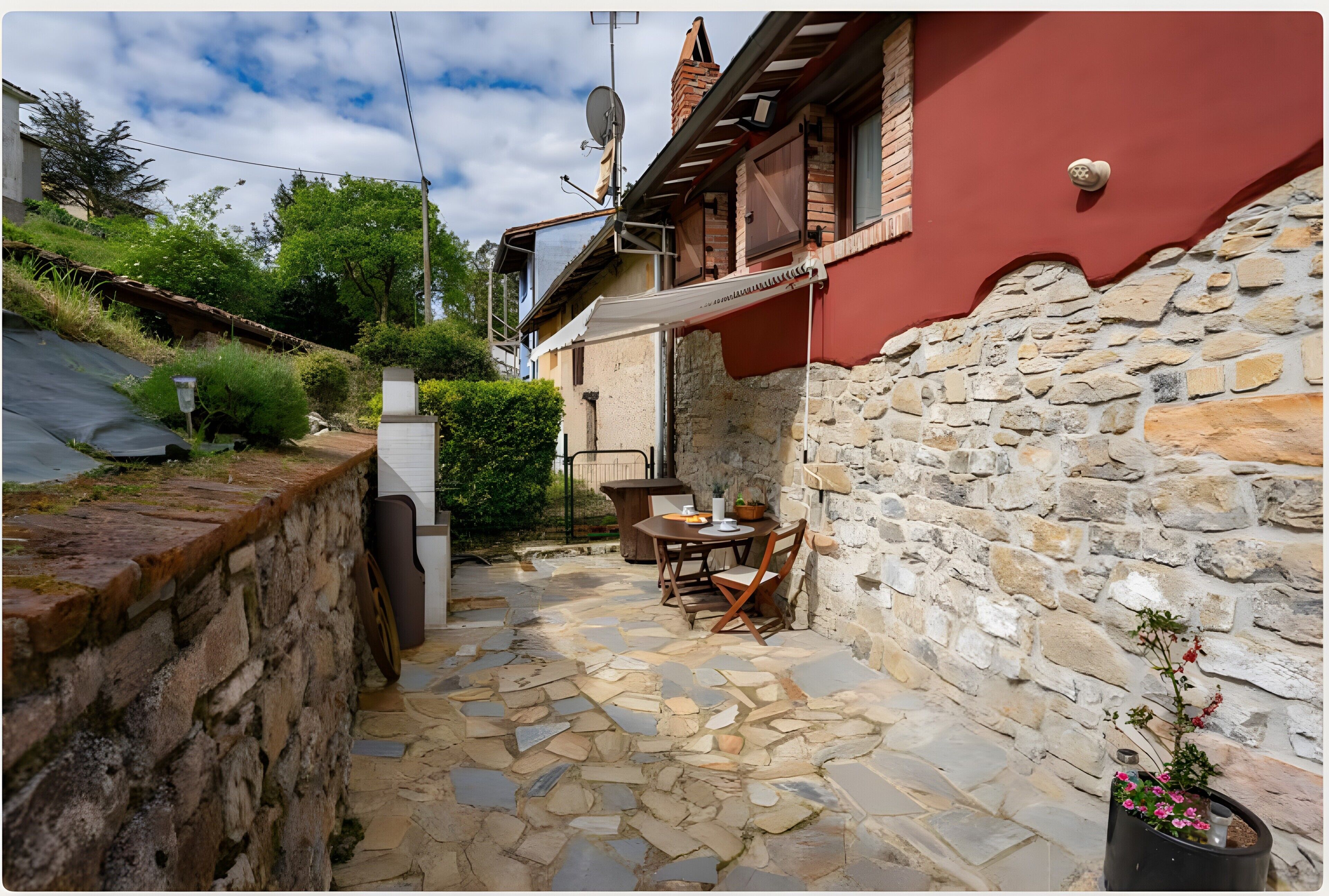 Un patio al aire libre con suelo de piedra, una mesa, sillas, una planta en maceta y un edificio rojo con techo inclinado.