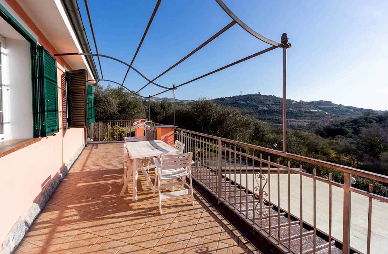 A balcony with a table and chairs, a railing, and a view of a hilly landscape.