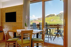 A dining area with wooden furniture, a flat-screen TV mounted on a wooden wall, and a balcony with a scenic view of a green landscape. - Holiday Apartment 'Baumannhof Apartment Aria' with Mountain View, Shared Terrace and Wi-Fi (Funes)