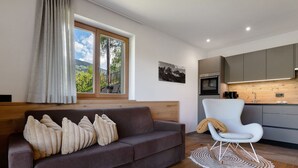 A modern living room with a sofa, a chair, a wooden floor, a window with a view, and a kitchen area. - Holiday Apartment 'Baumannhof Apartment Tera' with Mountain View, Shared Terrace and Wi-Fi (Funes)