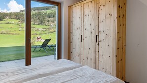 A bedroom with a large wooden wardrobe, a bed, and a sliding glass door leading to a balcony with a lawn and a chair. - Holiday Apartment 'Baumannhof Apartment Tera' with Mountain View, Shared Terrace and Wi-Fi (Funes)
