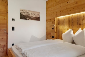 A bedroom with a wooden headboard, a bed with white bedding, a mountain landscape painting on the wall, and a thermostat.