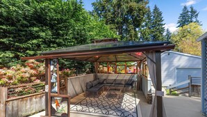 Un patio couvert doté d’un banc incurvé et d’une structure en verre, entouré d’une végétation luxuriante et d’une clôture en bois.