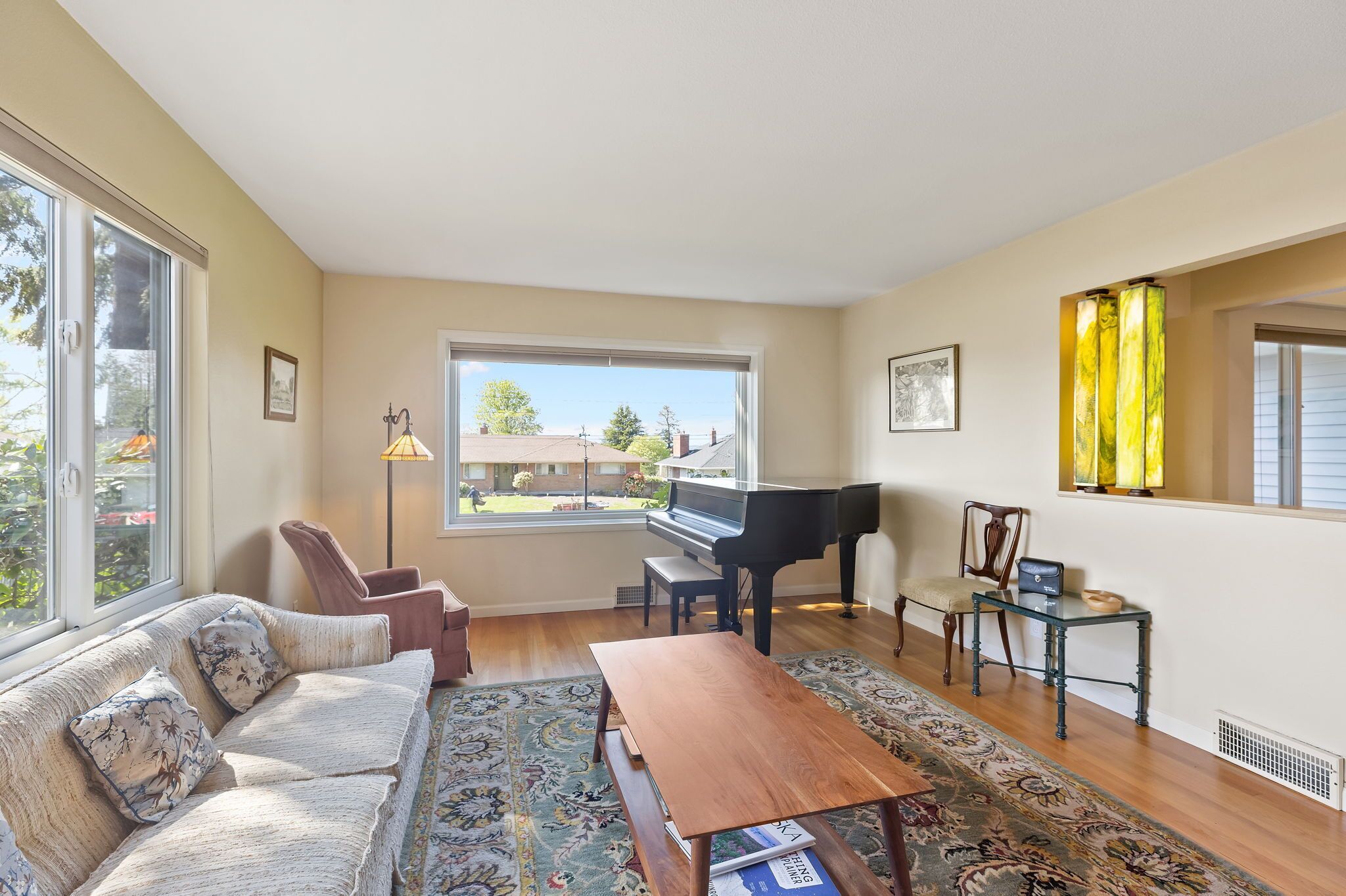 Un salon comprenant un canapé, un piano, une table basse et une vue sur une maison, visible à travers une grande fenêtre.