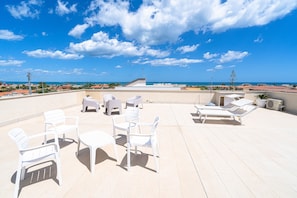 A rooftop terrace with white plastic chairs and tables, a view of the sea, and a clear sky.