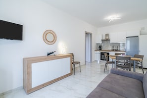 A modern living room with a wooden reception desk, a flat-screen TV mounted on the wall, a round mirror, and a dining area with a kitchen in the background.